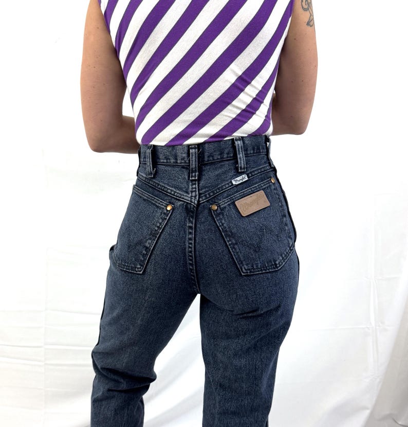 Puede incluir: Vaqueros Wrangler de mezclilla oscura con cintura alta y parche de cuero. Los vaqueros tienen remaches color lat&oacute;n y una peque&ntilde;a etiqueta Wrangler. Se ve una camiseta sin mangas con rayas diagonales moradas y blancas.