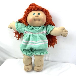 May include: A vintage doll with a round face, green eyes, and red yarn hair. The doll wears a mint green knit outfit with pink stripes and a floral trim. The doll's arms are outstretched, and it has a soft, plush body.
