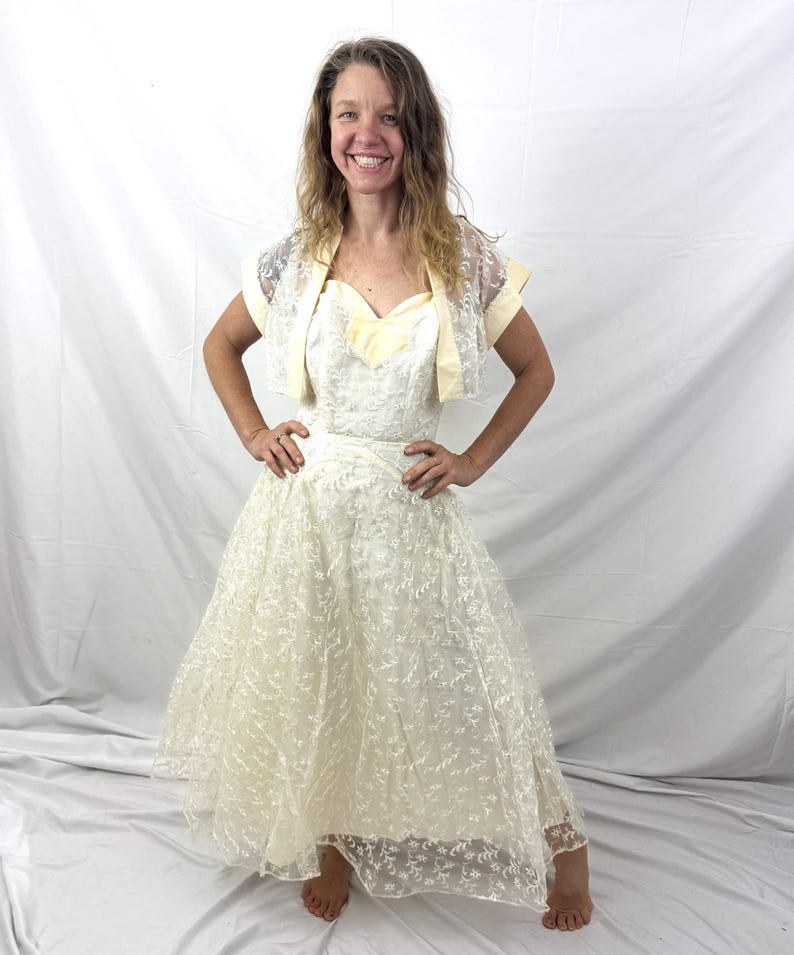 May include: A vintage ivory wedding dress with a sweetheart neckline and a full skirt. The dress is paired with a matching lace bolero jacket. The dress features delicate floral embroidery throughout. The bolero has short sleeves and a lace overlay.