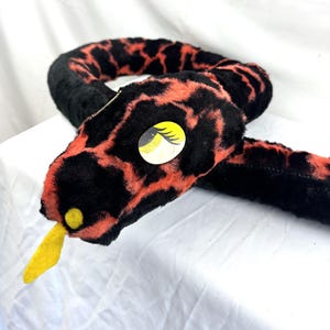 Puede incluir: Un juguete de serpiente de peluche con una textura suave y difusa con un patrón moteado en negro y naranja. La serpiente tiene una lengua de fieltro amarilla, una mancha amarilla en la nariz y un ojo grande y ovalado con iris amarillo y pestañas negras.