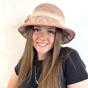 May include: A pink and beige cloche hat with a wide brim. The hat features a floral pattern, a cream-colored band, and a decorative accent on the side. The brim is trimmed with a pink rope-like material.