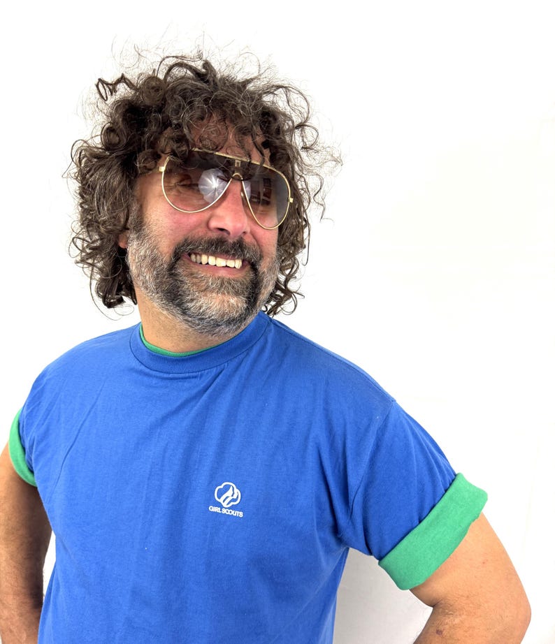 Puede incluir: Una camiseta azul con ribete verde y mangas enrolladas. La camiseta presenta el logotipo de las Girl Scouts en la parte delantera. La persona de la imagen lleva gafas de sol de aviador con montura dorada.