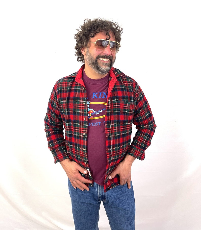 May include: A man wearing a red and green plaid flannel shirt over a maroon t-shirt with the text "King's Island" and a graphic of a guitar. He is wearing blue jeans and sunglasses.