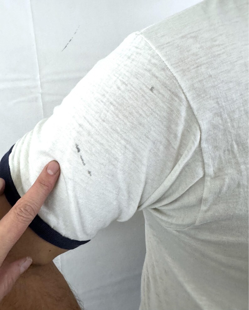 May include: Cream-colored short-sleeve shirt with navy blue trim on the sleeve. The shirt is made of a soft, textured fabric. The image shows a close-up of the sleeve and part of the arm.