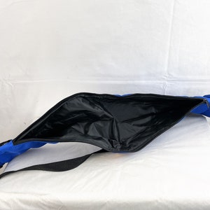 May include: A bright blue elongated bag with a wide, open top featuring a black zipper closure. The interior is lined with a smooth black material. A black adjustable strap is attached to the exterior of the blue fabric bag.