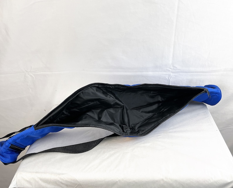 May include: A bright blue elongated bag with a wide, open top featuring a black zipper closure. The interior is lined with a smooth black material. A black adjustable strap is attached to the exterior of the blue fabric bag.