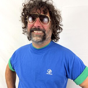 Puede incluir: Una camiseta azul con ribetes verdes y mangas enrolladas. La camiseta presenta el logotipo de las Girl Scouts en el pecho. La persona de la imagen lleva gafas de sol de aviador con montura dorada.