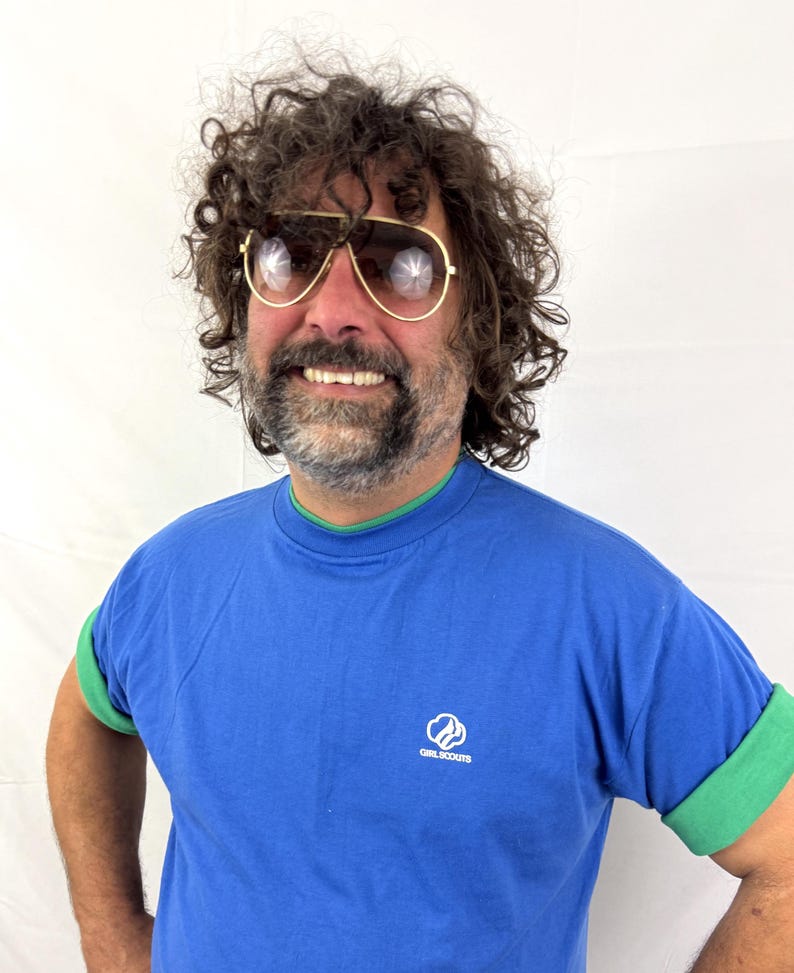 Puede incluir: Una camiseta azul con ribetes verdes y mangas enrolladas. La camiseta presenta el logotipo de las Girl Scouts en el pecho. La persona de la imagen lleva gafas de sol de aviador con montura dorada.
