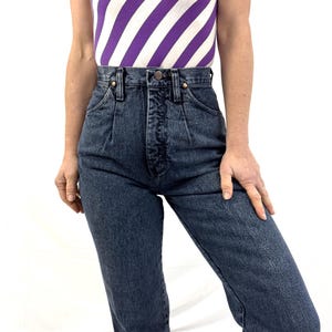Puede incluir: Vaqueros de mezclilla de cintura alta, de lavado oscuro, con cierre de bot&oacute;n y pliegues delanteros. Los vaqueros se combinan con una camiseta sin mangas con rayas diagonales moradas y blancas. El conjunto est&aacute; siendo usado.