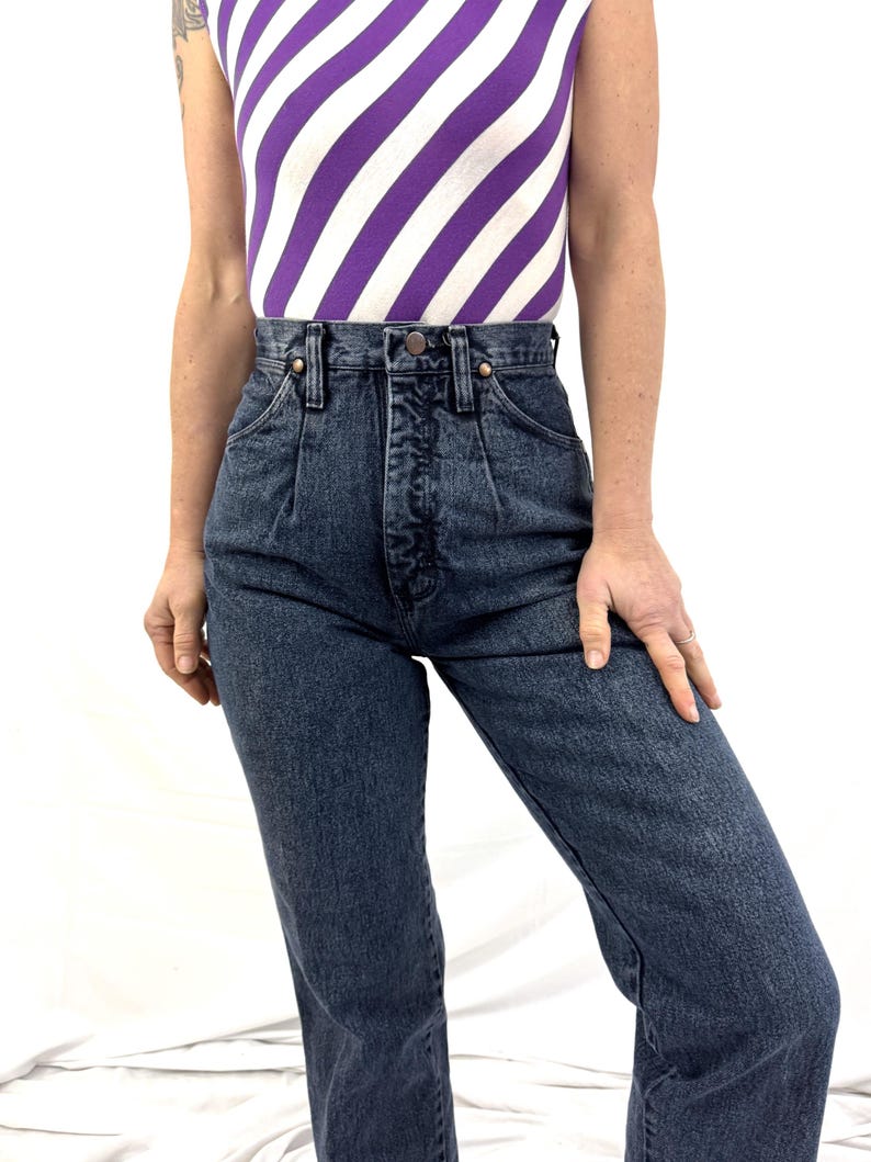 Puede incluir: Vaqueros de mezclilla de cintura alta, de lavado oscuro, con cierre de bot&oacute;n y pliegues delanteros. Los vaqueros se combinan con una camiseta sin mangas con rayas diagonales moradas y blancas. El conjunto est&aacute; siendo usado.