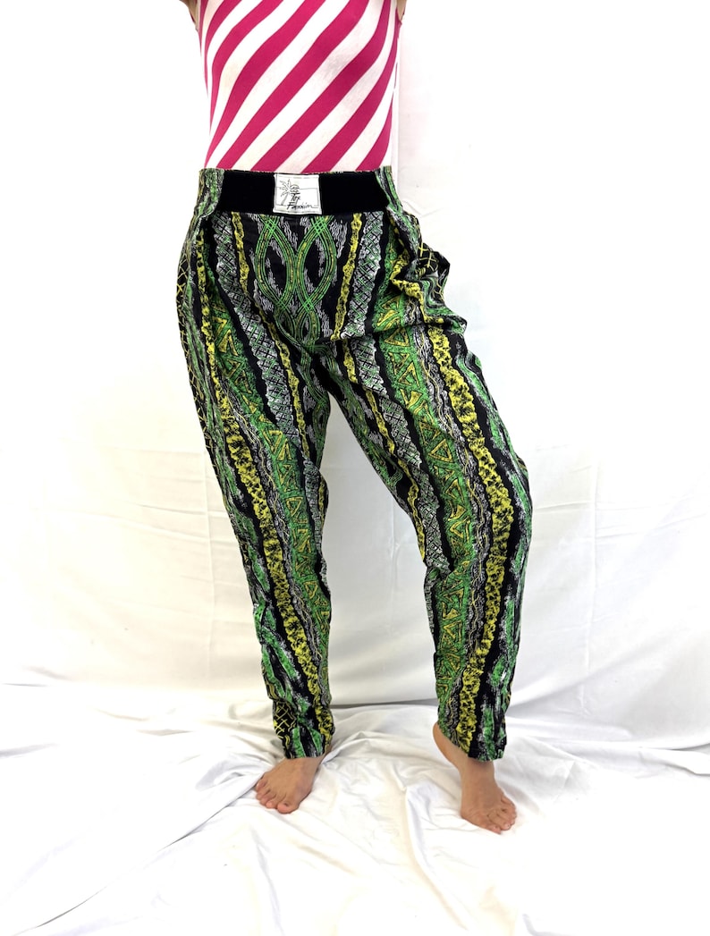 May include: Green, yellow, and black patterned pants with a black waistband. The pants have a unique design with vertical stripes and geometric shapes. The model is also wearing a pink and white striped top.
