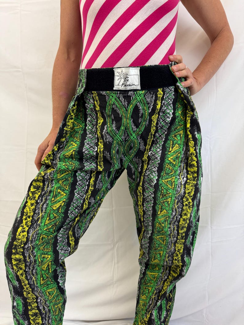 May include: Green, yellow, and black patterned pants with a wide black waistband. The pants feature a repeating geometric design. The waistband has a tag with text. The top is a pink and white striped tank top.