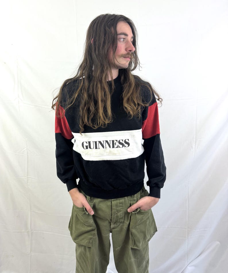 May include: Black sweatshirt with red shoulder accents and a white panel across the chest. The word "GUINNESS" is printed in black on the white panel. The person is wearing olive green cargo pants.