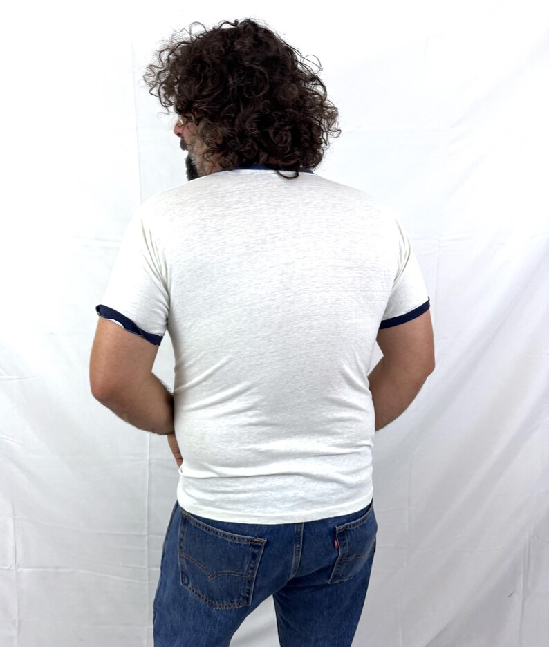 May include: A white ringer t-shirt with navy blue trim on the collar and sleeves. The shirt is paired with blue denim jeans. The person is facing away from the camera.