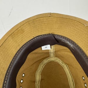 May include: Tan safari hat with a brown leather band and eyelets. The hat has a medium size tag inside. The brim is stitched and the hat is made of a durable material.