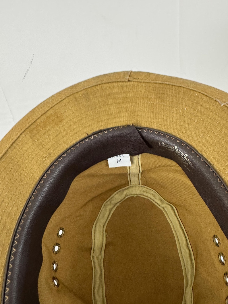 May include: Tan safari hat with a brown leather band and eyelets. The hat has a medium size tag inside. The brim is stitched and the hat is made of a durable material.