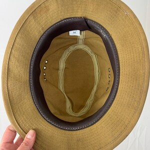 May include: Tan safari hat with a brown leather band and interior trim. The hat has a wide brim and a fabric sweatband. The interior features a size tag marked "M".