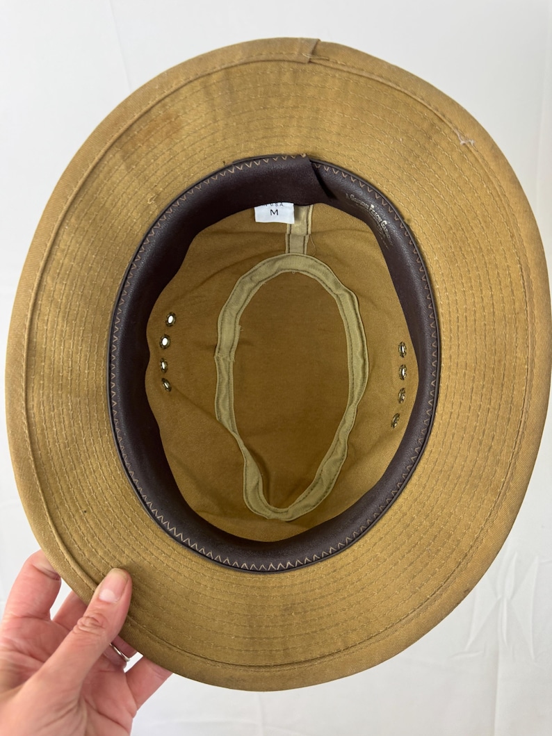 May include: Tan safari hat with a brown leather band and interior trim. The hat has a wide brim and a fabric sweatband. The interior features a size tag marked "M".
