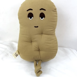 May include: A tan-colored, bean-shaped plush pillow with embroidered eyes, eyebrows, and a mouth. The pillow has a twisted hand-like appendage and a small tag. The pillow is on a white surface.