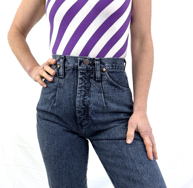 Puede incluir: Vaqueros de mezclilla de lavado oscuro de cintura alta con cierre de bot&oacute;n y cremallera. Los vaqueros tienen un dise&ntilde;o cl&aacute;sico de cinco bolsillos y una parte delantera plisada. El modelo lleva una camiseta sin mangas a rayas moradas y blancas.