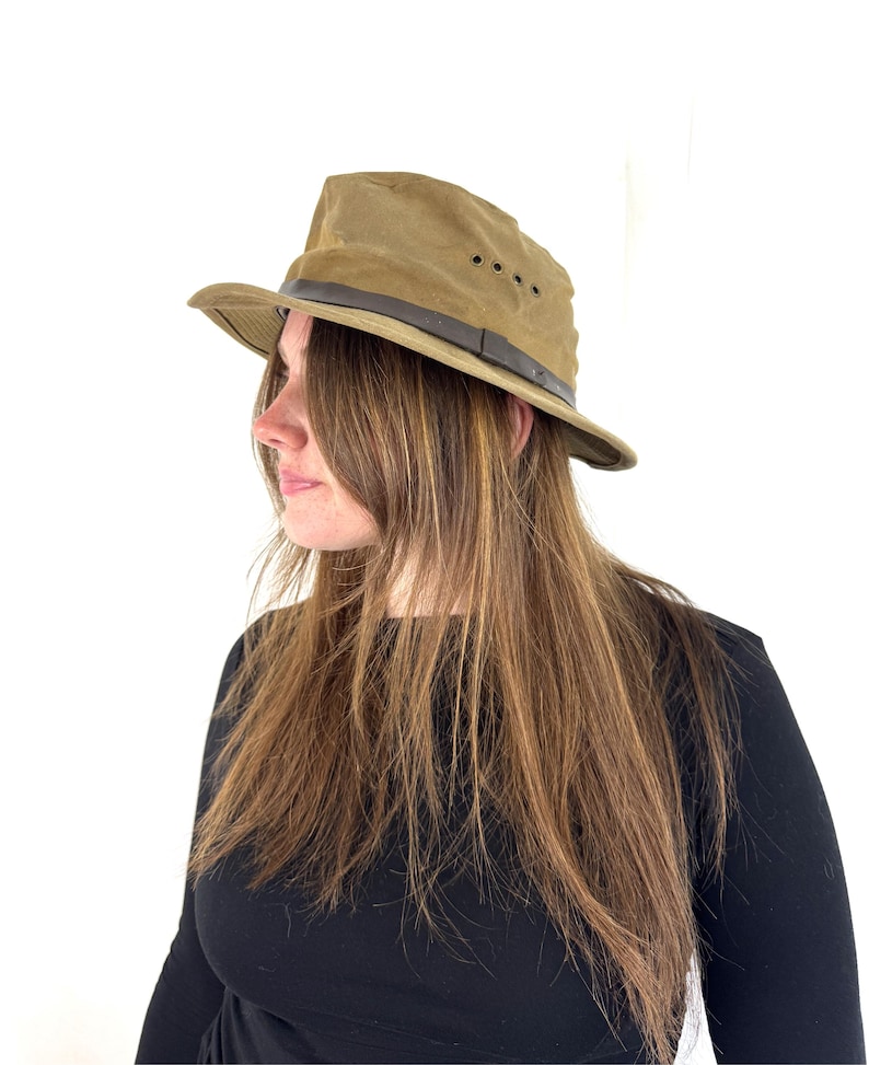May include: A khaki-colored, wide-brimmed hat with a dark brown leather band and eyelets. The hat has a structured crown and a slightly curved brim. The hat is designed for outdoor wear and protection from the sun.