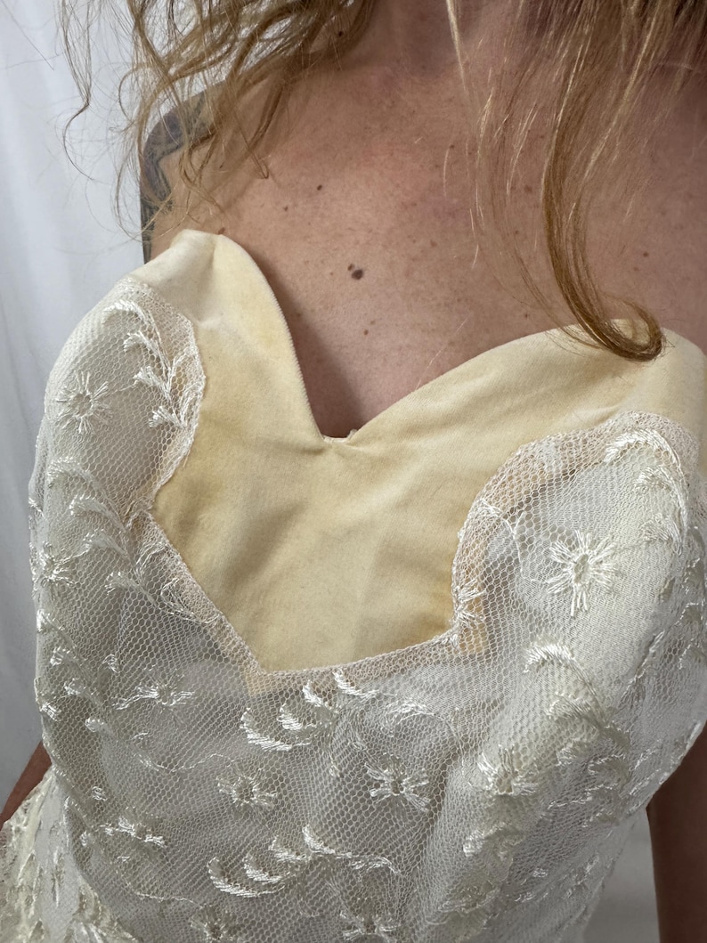 Puede incluir: Cuerpo de vestido sin tirantes color crema con escote coraz&oacute;n. El corpi&ntilde;o est&aacute; hecho de una tela suave de color amarillo claro, cubierta con delicado encaje blanco con motivos florales. El encaje se extiende sobre los hombros.