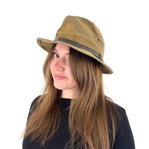May include: A tan fedora-style hat with a dark brown leather band. The hat has a slightly distressed appearance and a classic brim. The hat is made of a durable material and is suitable for outdoor wear.