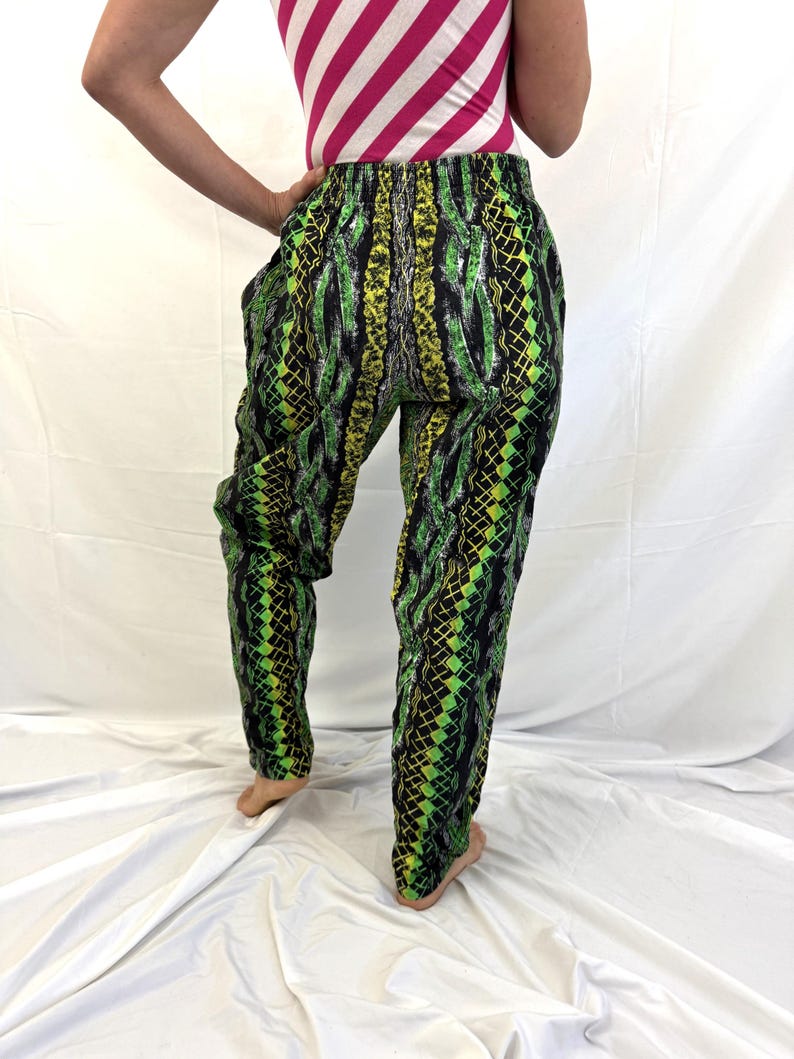 May include: Green, black, and yellow patterned pants. The pants have a snake-like design and a loose fit. The person in the image is wearing a pink and white striped top.