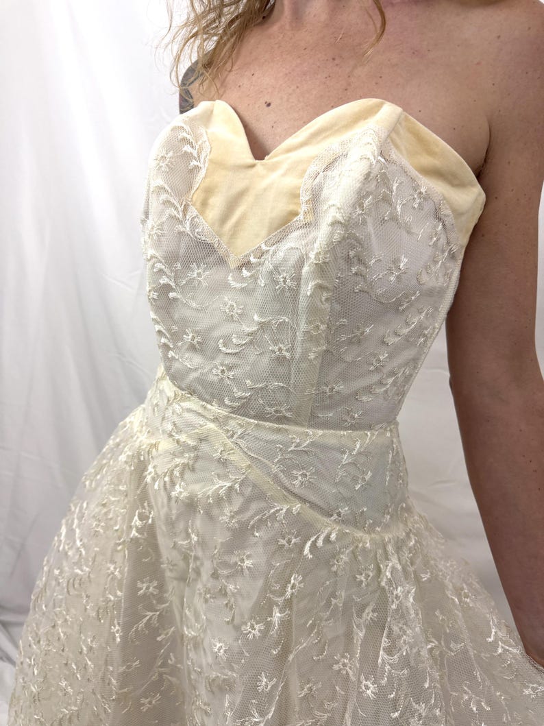 Puede incluir: Vestido vintage marfil sin tirantes con escote coraz&oacute;n. El corpi&ntilde;o presenta un ribete de terciopelo crema y est&aacute; cubierto con delicado encaje floral. La falda se ensancha, creando una silueta cl&aacute;sica.
