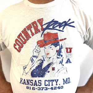 Puede incluir: Camiseta gris claro con un gr&aacute;fico que presenta a una mujer con un sombrero de vaquero rojo, las palabras "Country Rock" y "Kansas City, MO." El dise&ntilde;o incluye acentos rojos, blancos y azules, y el texto "USA."