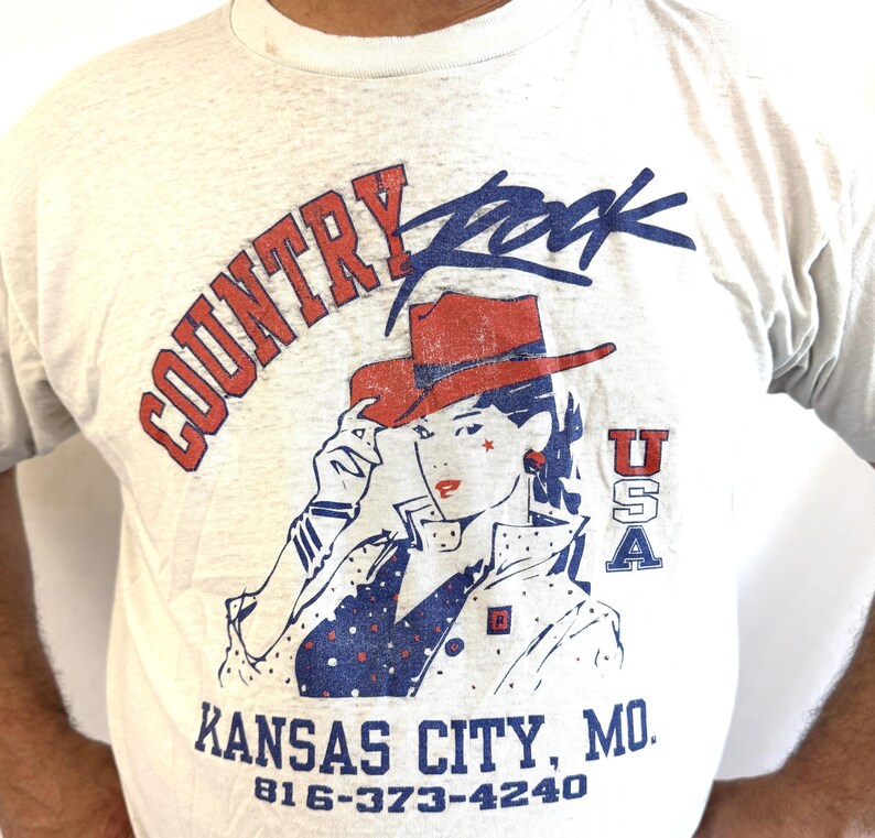 Puede incluir: Camiseta gris claro con un gr&aacute;fico que presenta a una mujer con un sombrero de vaquero rojo, las palabras "Country Rock" y "Kansas City, MO." El dise&ntilde;o incluye acentos rojos, blancos y azules, y el texto "USA."
