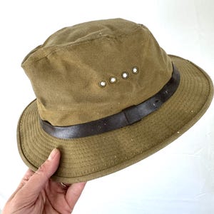 May include: Olive green hat with a brown leather band. The hat has a brim and four eyelets on the side. The hat is made of a durable fabric.