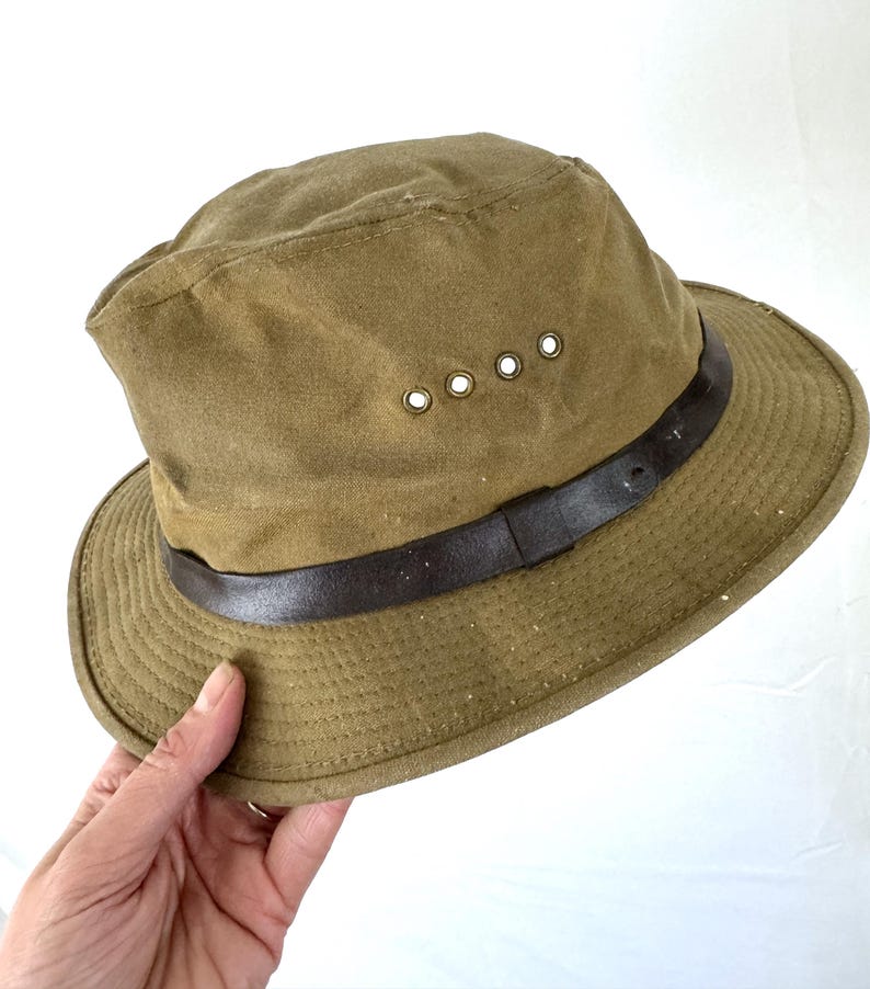 May include: Olive green hat with a brown leather band. The hat has a brim and four eyelets on the side. The hat is made of a durable fabric.