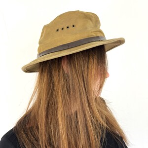 May include: A khaki-colored safari hat with a dark brown leather band and four ventilation eyelets. The hat has a wide brim and a slightly worn appearance. The hat is a classic outdoor accessory.