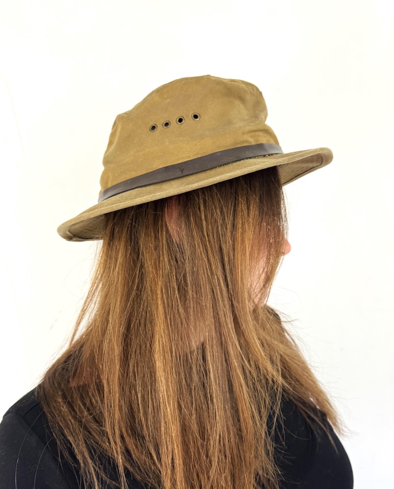 May include: A khaki-colored safari hat with a dark brown leather band and four ventilation eyelets. The hat has a wide brim and a slightly worn appearance. The hat is a classic outdoor accessory.