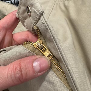 Puede incluir: Primer plano de un par de pantalones beige con una cremallera dorada y un bot&oacute;n marr&oacute;n. La cremallera tiene la palabra "YKK" impresa en ella.
