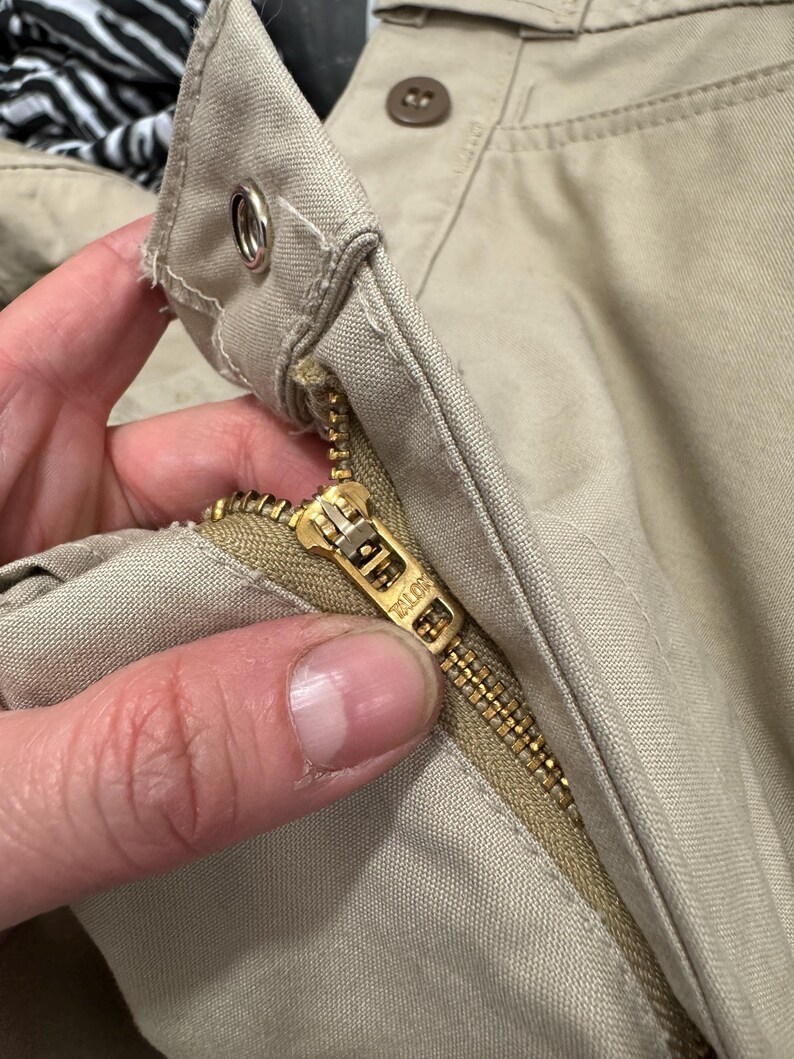 Puede incluir: Primer plano de un par de pantalones beige con una cremallera dorada y un bot&oacute;n marr&oacute;n. La cremallera tiene la palabra "YKK" impresa en ella.