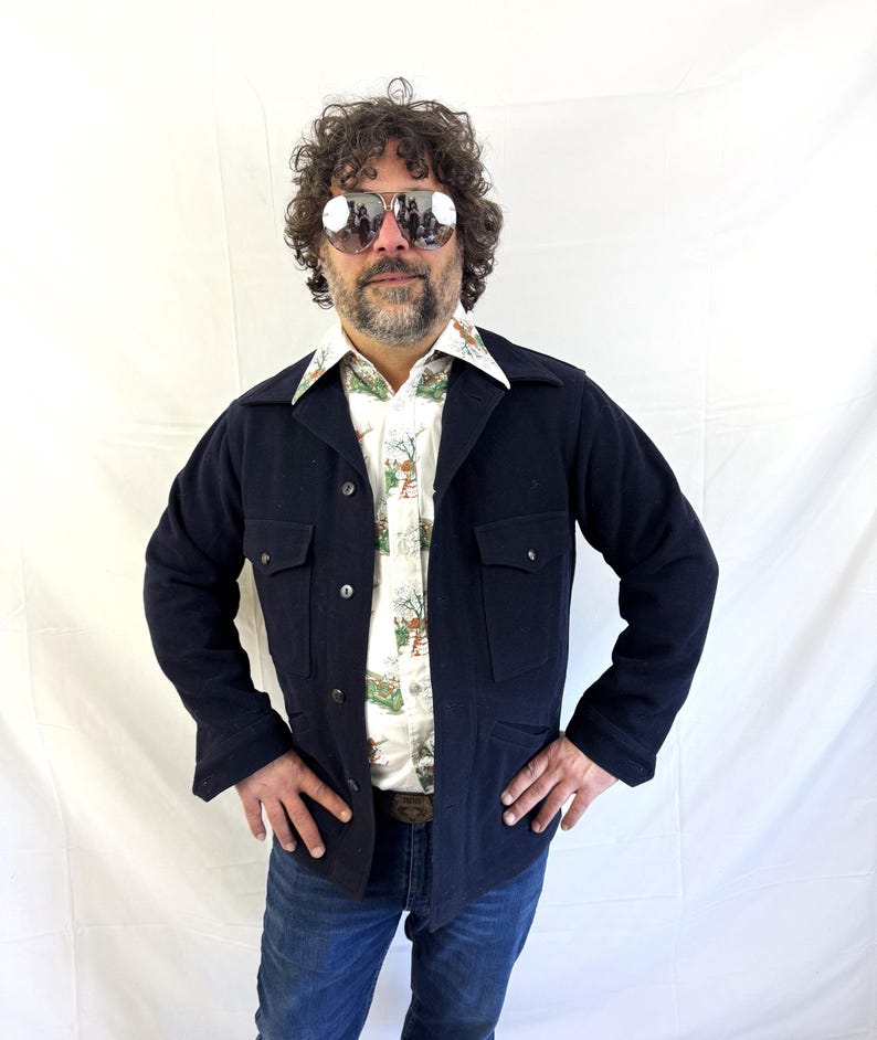 Puede incluir: Una chaqueta azul marino con dos bolsillos en el pecho y dos bolsillos inferiores. La chaqueta se usa sobre una camisa con botones estampada y jeans azules. La persona lleva gafas de sol de aviador.