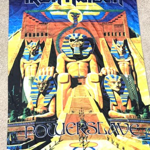 Può includere: Un arazzo nero, blu e giallo di Iron Maiden Powerslave. L'arazzo presenta il logo della band in alto, con un'illustrazione di un faraone e figure egizie. La parola "Powerslave" è scritta in blu in basso.