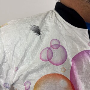 May include: White bomber jacket with a black collar and a feather and bubble design. The jacket features colorful bubbles in orange, pink, and purple. The feather is black and grey.