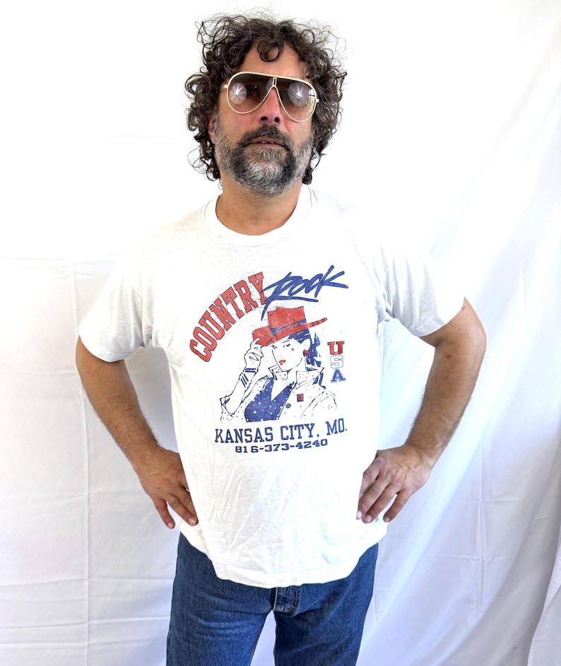 Puede incluir: Camiseta blanca con un gr&aacute;fico que dice "Country Rock" en rojo y azul, con una mujer con sombrero de vaquero. La camiseta tambi&eacute;n muestra "Kansas City, MO" y un n&uacute;mero de tel&eacute;fono. La persona lleva vaqueros azules y gafas de sol de aviador.