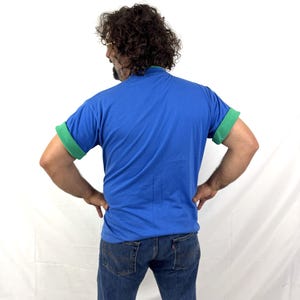 Puede incluir: Una camiseta azul con mangas verdes enrolladas y cuello verde. La persona de la imagen lleva vaqueros azules. El fondo es blanco.