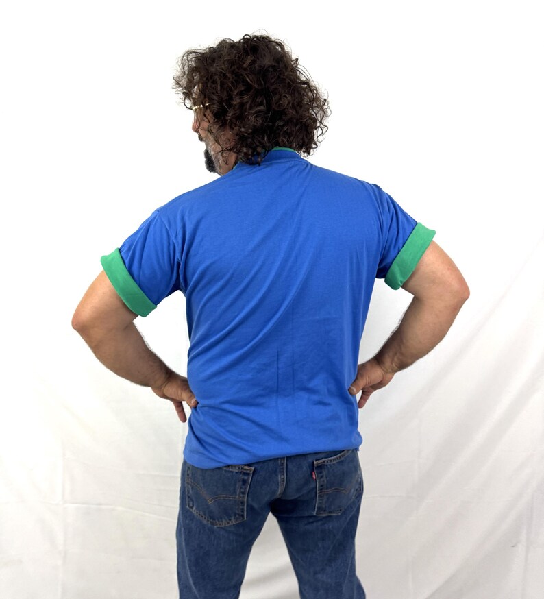 Puede incluir: Una camiseta azul con mangas verdes enrolladas y cuello verde. La persona de la imagen lleva vaqueros azules. El fondo es blanco.