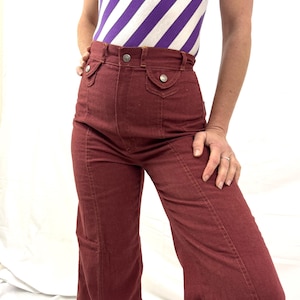 May include: A pair of high-waisted, flared burgundy pants with a button and pocket detail. The pants are paired with a sleeveless top with purple and white diagonal stripes. The pants have a vintage style.