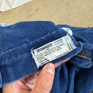 K&ouml;nnte beinhalten: Ein blaues Wrangler-Jeans-Etikett aus Denim mit wei&szlig;em Text. Das Etikett lautet "Machine wash warm with like colors, only non-chlorine bleach when needed, tumble dry low, remove promptly. Made in USA 100% Cotton 22MW3BA 7/8 x 34 0 94654 52504 5"
