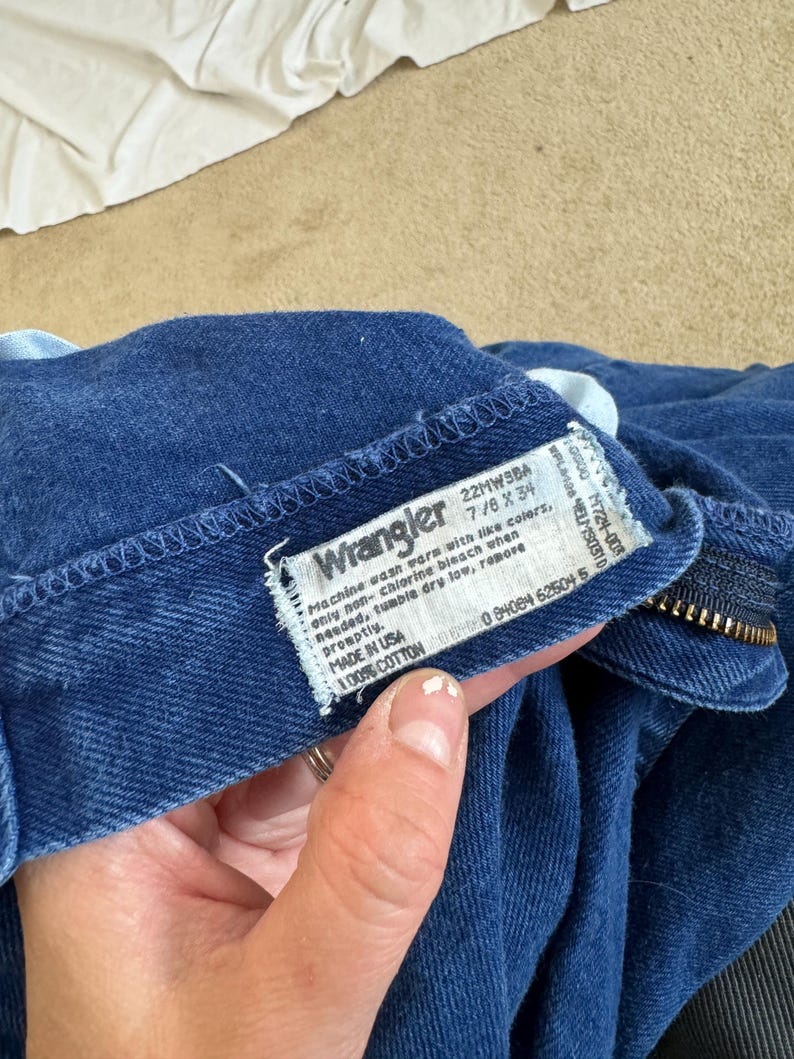 K&ouml;nnte beinhalten: Ein blaues Wrangler-Jeans-Etikett aus Denim mit wei&szlig;em Text. Das Etikett lautet "Machine wash warm with like colors, only non-chlorine bleach when needed, tumble dry low, remove promptly. Made in USA 100% Cotton 22MW3BA 7/8 x 34 0 94654 52504 5"