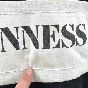 May include: Close-up of a black garment with a white band across the chest. The word "GUINNESS" is printed in large, bold, black letters on the white band. The fabric appears to be slightly worn.