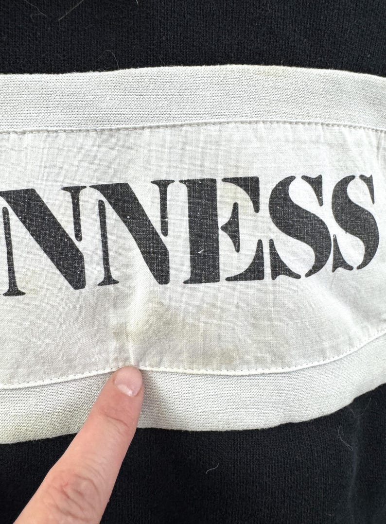 May include: Close-up of a black garment with a white band across the chest. The word "GUINNESS" is printed in large, bold, black letters on the white band. The fabric appears to be slightly worn.