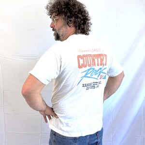 Puede incluir: Camiseta blanca con el texto "I Learned to Dance in Country Rock USA" en rojo y azul. La camiseta tambi&eacute;n presenta el texto "Kansas City" y un n&uacute;mero de tel&eacute;fono. La persona lleva vaqueros azules.