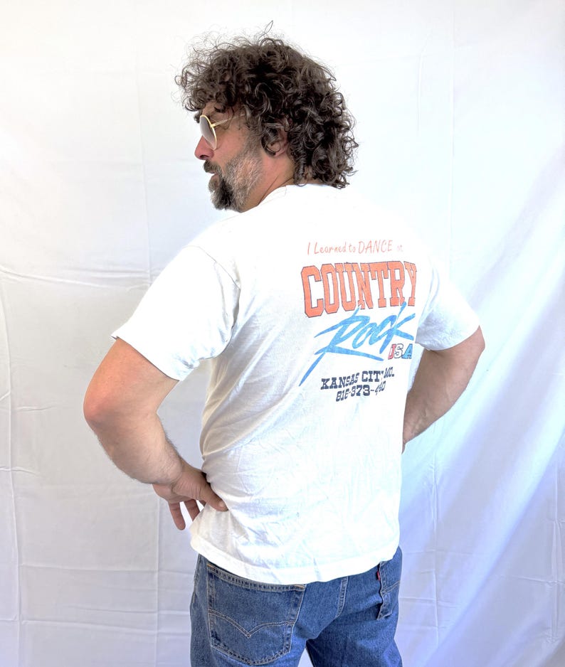 Puede incluir: Camiseta blanca con el texto "I Learned to Dance in Country Rock USA" en rojo y azul. La camiseta tambi&eacute;n presenta el texto "Kansas City" y un n&uacute;mero de tel&eacute;fono. La persona lleva vaqueros azules.